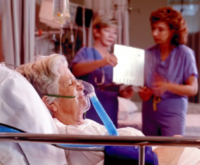 Elderly woman in an oxygen mask in hospital bed while medical professionals check her chart