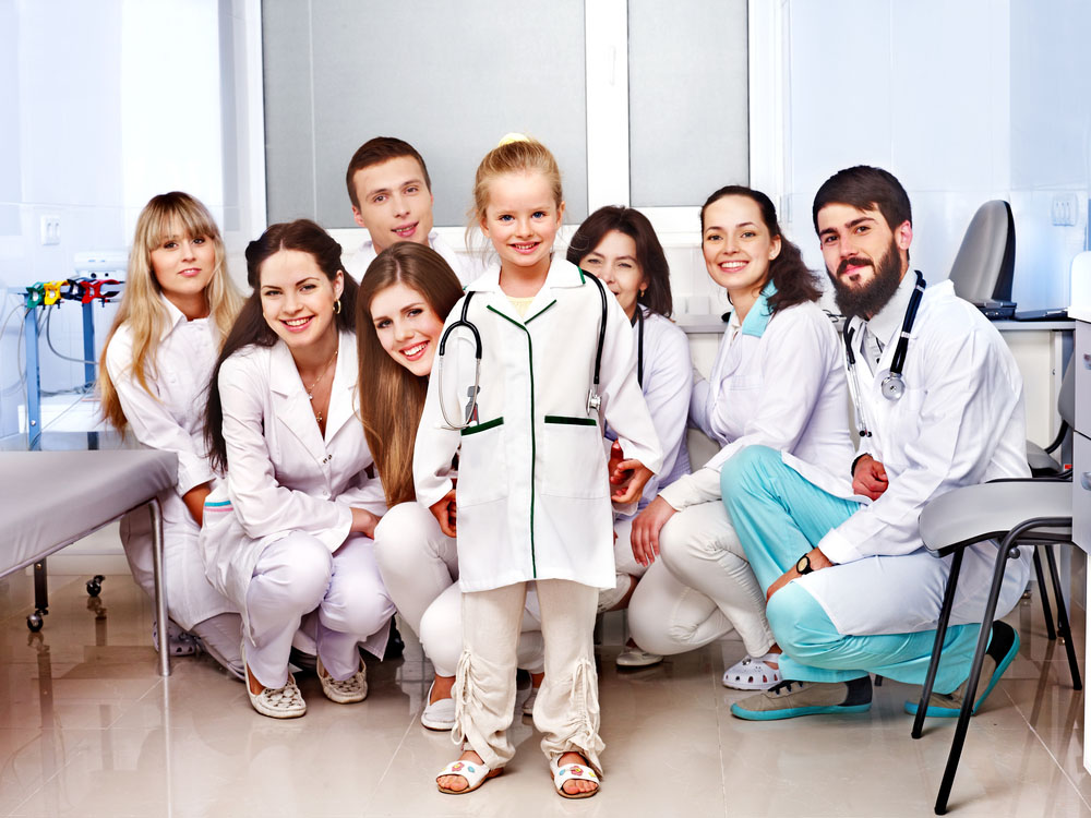Little girl wearing a doctor's coat and stethoscope with medical professionals surrounding her, posing for the camera